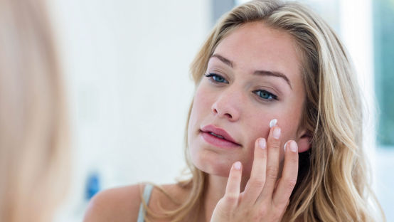 Smiling pretty woman applying cream on her face in bathroom